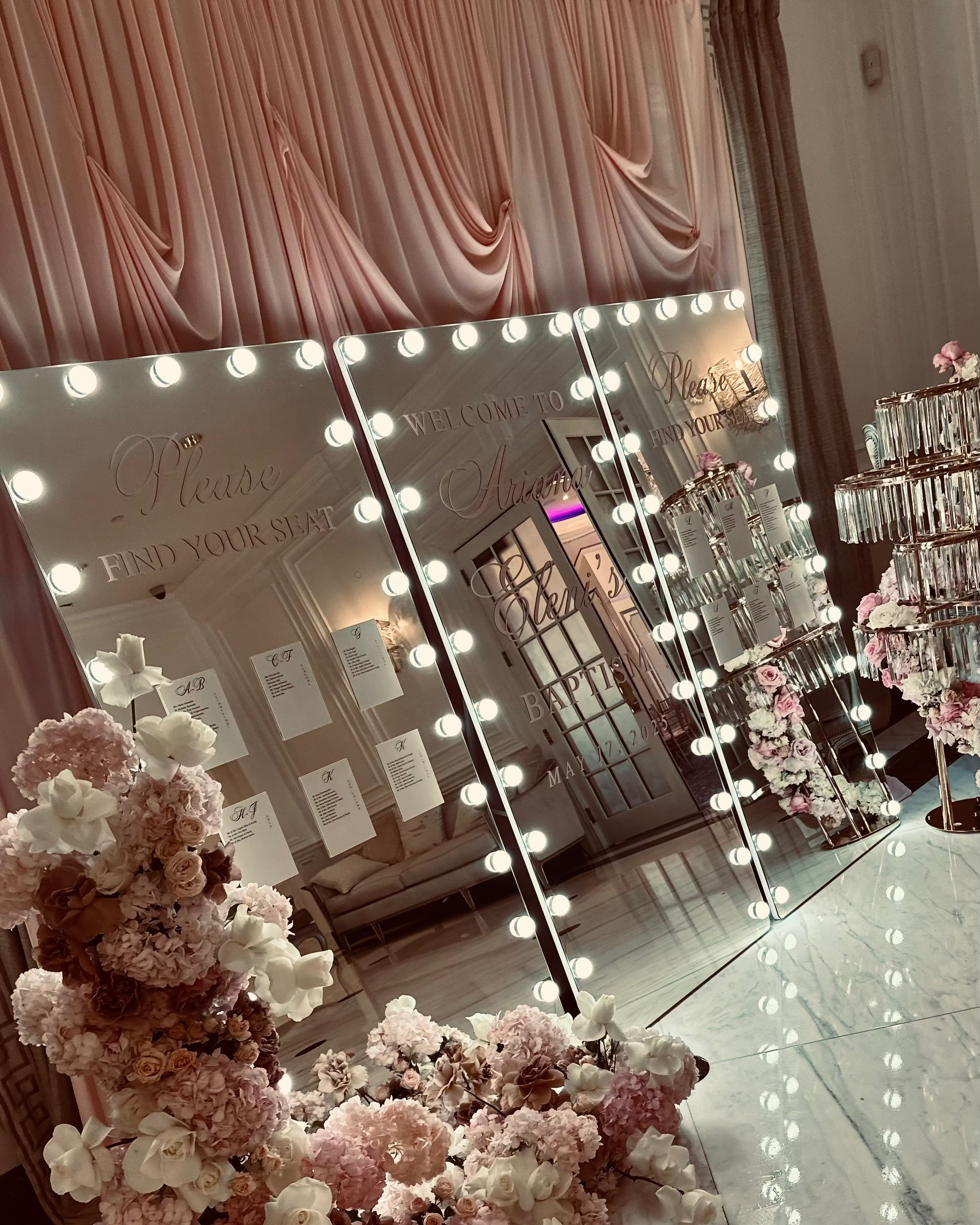 Decorative hollywood mirror welcome sign with lights surrounded by floral arrangements and pink drapes. 